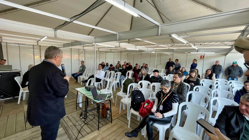A imagem mostra uma pessoa, de costas, falando para uma plateia usando um microfone. A pessoa está em pé, usando um terno preto e tênis brancos, e está posicionada à esquerda da imagem. Diante dela, há um pequeno balcão com um laptop e alguns papéis. A plateia é composta por várias pessoas sentadas em cadeiras brancas de plástico, dentro de uma estrutura coberta por uma tenda com armação metálica. Algumas pessoas estão prestando atenção ao palestrante, enquanto outras parecem estar olhando para seus dispositivos móveis ou conversando entre si. No fundo, há mais algumas pessoas em pé, próximas às paredes laterais da tenda.