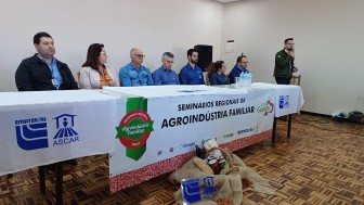 Autoridades sentadas em mesa com toalha branca e com uma faixa escrita "Seminários Regionais de Agroindústria Familiar". Enquanto isso, o diretor substituto do Departamento de Agroindústria Familiar da SDR, fala no evento.