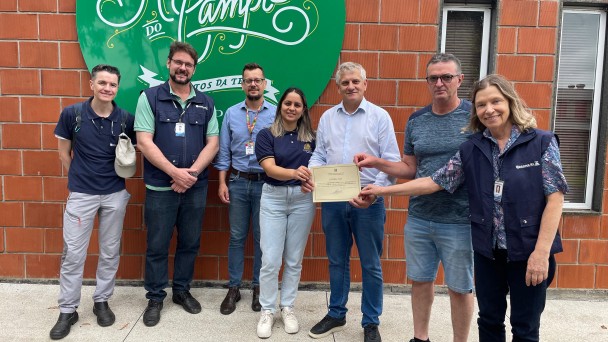 Sete pessoas posam para a foto à frente de uma parede de tijolo à vista. Quatro delas seguram um certificado.