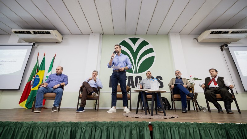 Governador fala, à frente, enquanto cinco autoridades estão sentadas no palco